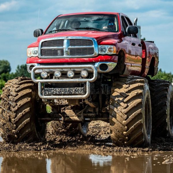 Na zdjęciu widać potężnego monster trucka w czerwonym kolorze, z wielkimi błotnistymi oponami, jadącego przez błotnisty teren pod błękitnym niebem.