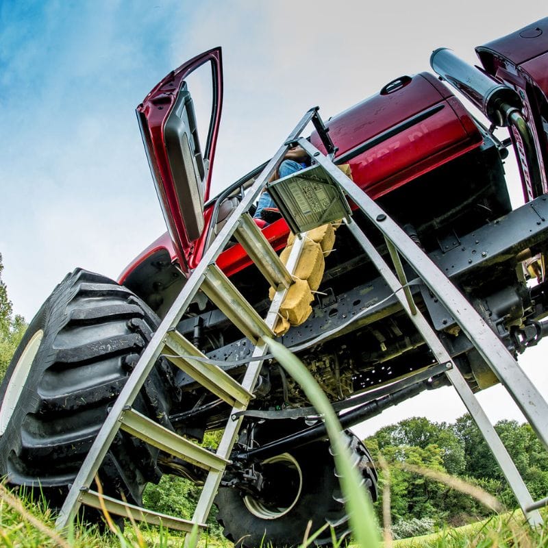 Widok od dołu na potężnego monster trucka z otwartymi drzwiami, przy którym stoi drabina, a całość otacza bujna zieleń.