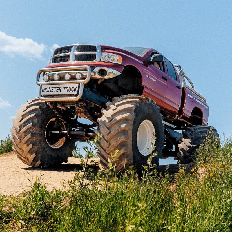 Na grafice widzimy imponujący monster truck na dużych kołach, który dumnie prezentuje się na tle błękitnego nieba i zielonej przyrody, będąc doskonałą atrakcją dla fanów motoryzacyjnych emocji.