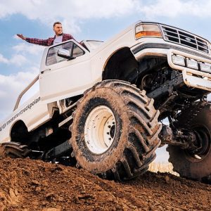 Mężczyzna w flanelowej koszuli z entuzjazmem wystaje przez okno potężnego monster trucka, który wspina się na stromej górce błota.