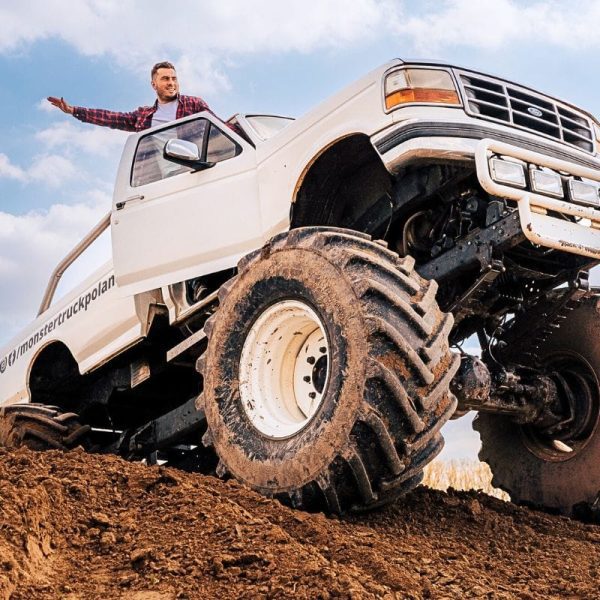 Mężczyzna w flanelowej koszuli z entuzjazmem wystaje przez okno potężnego monster trucka, który wspina się na stromej górce błota.