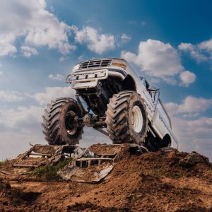 Ogromny monster truck w dramatycznym skoku nad starymi wrakami samochodów na tle nieba pełnego białych chmur.