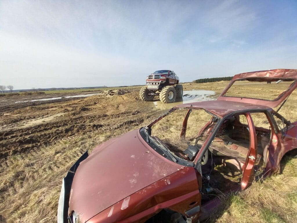 Na fotografii widać monster trucka na błotnistym terenie i wrak samochodu, co podkreśla emocje związane z atrakcjami terenowymi w Gassy pod Warszawą.