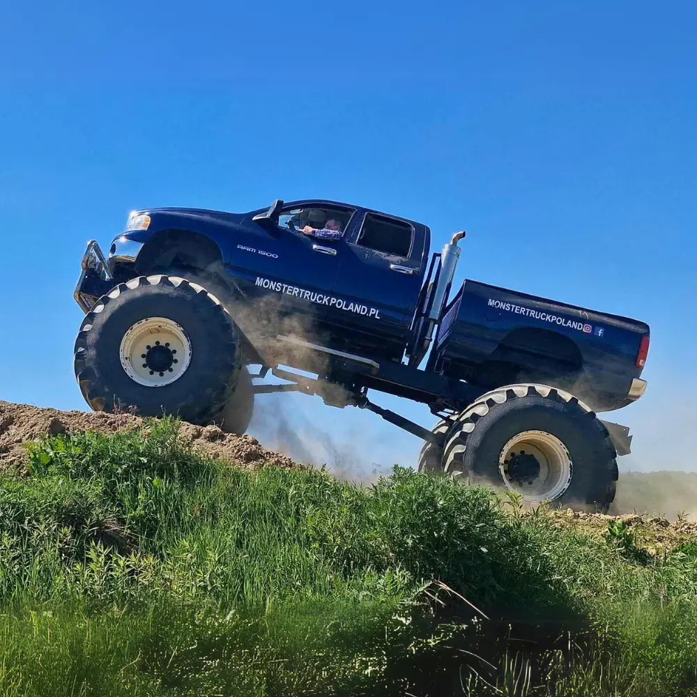 Na zdjęciu widać ogromnego, niebieskiego monster trucka wspinającego się po pagórku na tle błękitnego nieba, promującego atrakcje w Gassy pod Warszawą.