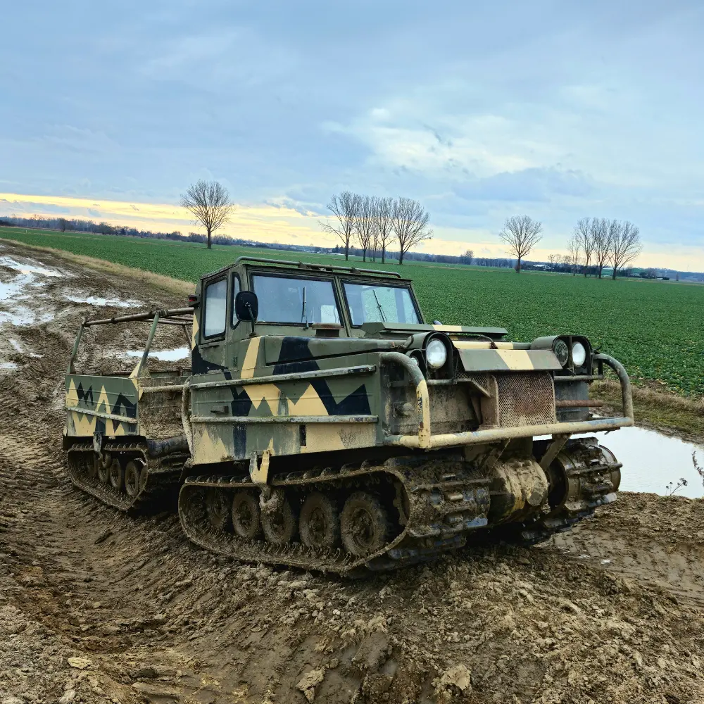 Pojazd terenowy o gąsienicach w kamuflażowych barwach stoi na błotnistej drodze z widokiem na pola i rząd drzew na horyzoncie.