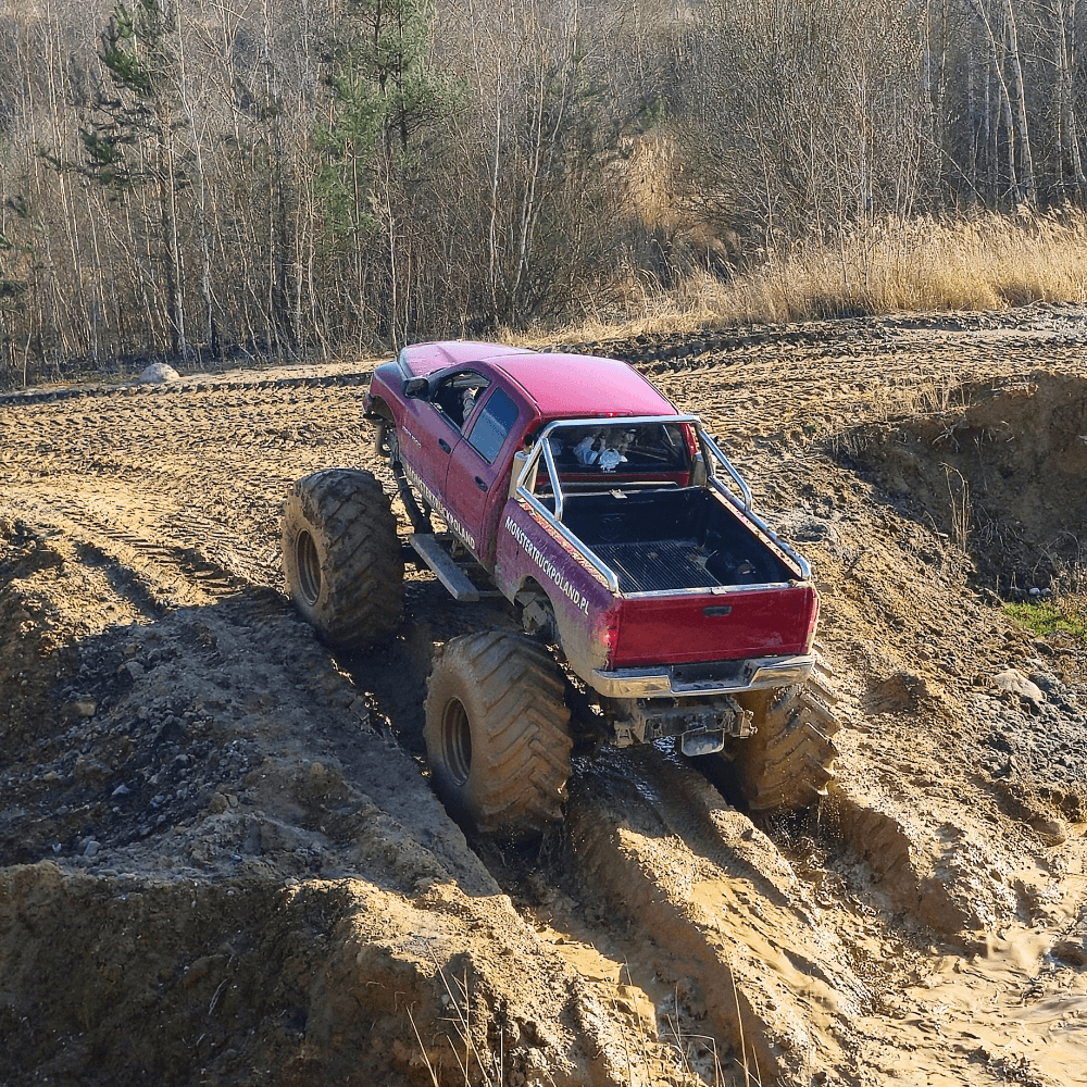 Różowy monster truck z masywnymi kołami przejeżdża przez błotnistą ścieżkę w lesie, oferując ekscytującą atrakcję terenową niedaleko Warszawy w Gassach.