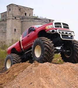 Potężny monster truck w intensywnej czerwieni wspina się na ziemisty nasyp, mając w tle monumentalny budynek, tworząc wrażenie ekscytującej przygody.