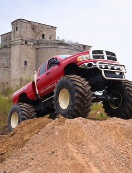 Potężny monster truck w intensywnej czerwieni wspina się na ziemisty nasyp, mając w tle monumentalny budynek, tworząc wrażenie ekscytującej przygody.