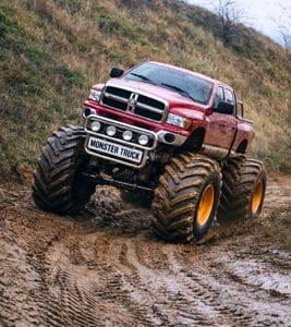 Czerwony monster truck z ogromnymi kołami przemierza błotnistą trasę w malowniczym otoczeniu.
